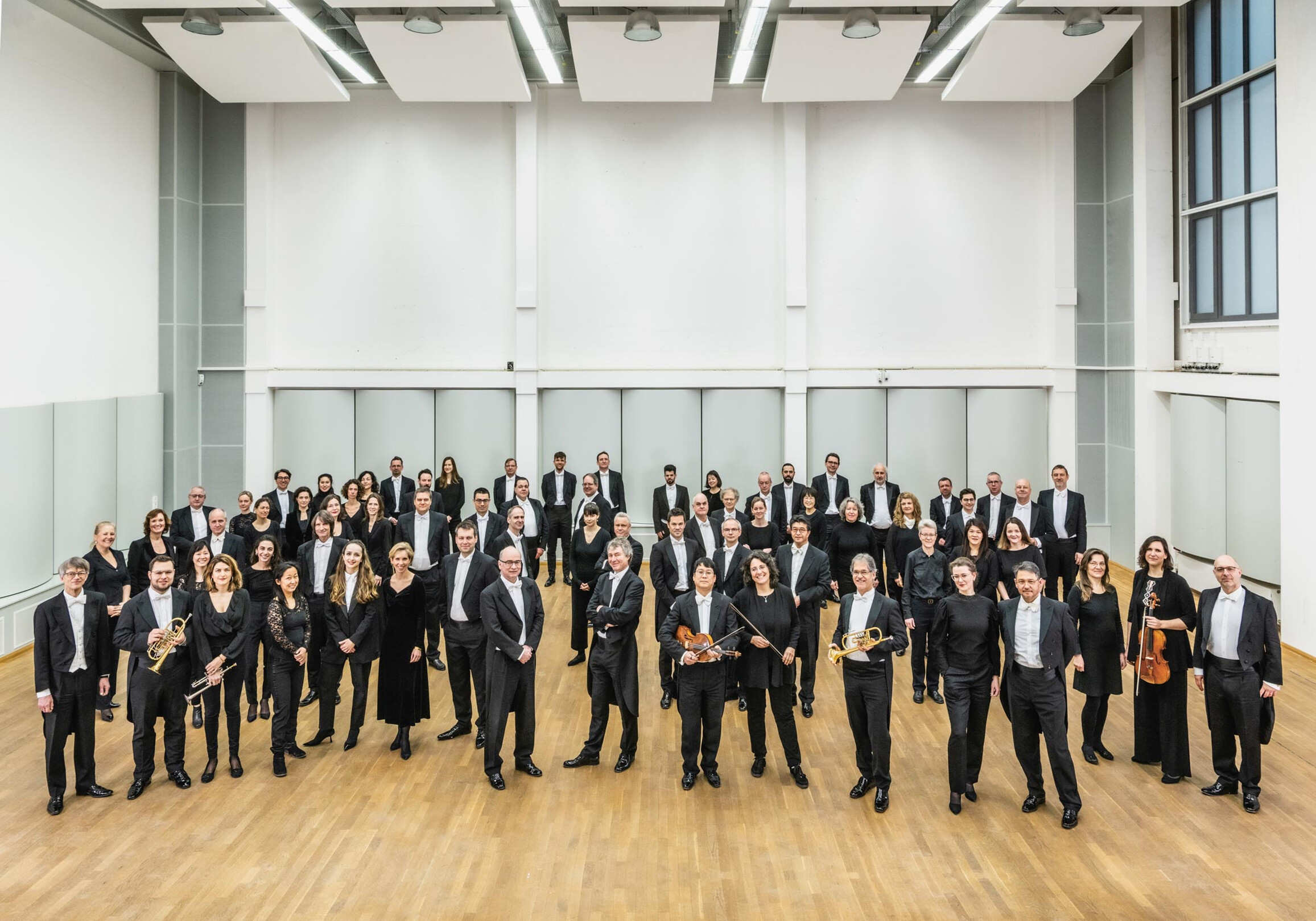 Orchestermusiker:nnen stehend im Probenraum, einige mit Instrumenten in der Hand